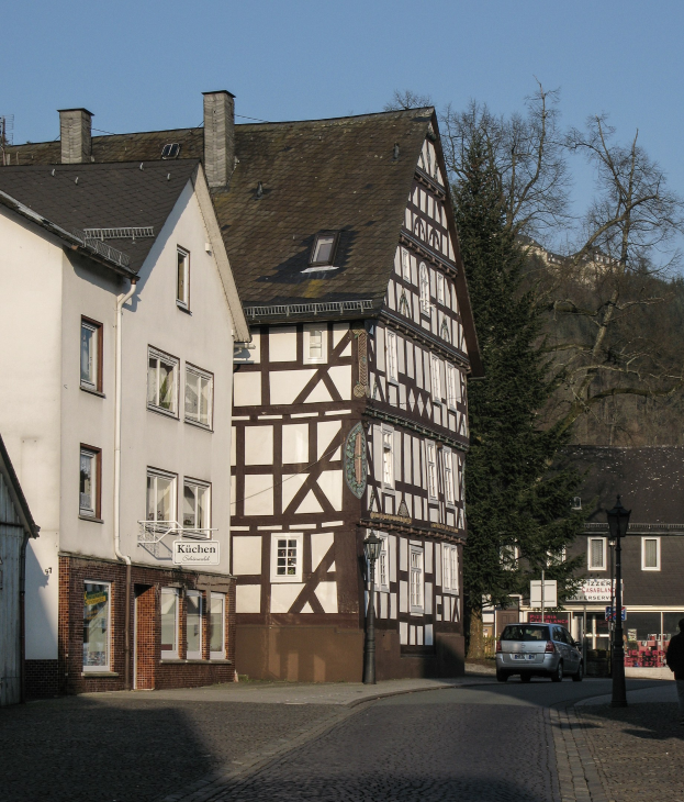 Half-timbered house in a small town surrounded by buildings, vehicles, pedestrians, trees, and street infrastructure under a clear blue sky.