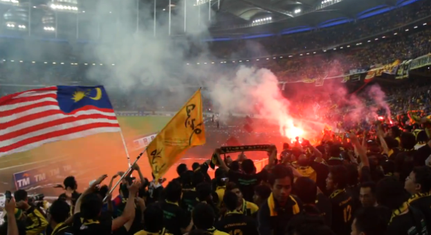 Eine große Menschenmenge in einem Stadion mit Fackeln, Fahnen und einer Kamera, mit Rauch und Schildern, die "Malaysia vs Malaysia Live Streaming" im Hintergrund zeigen.