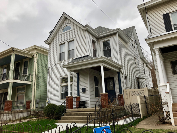 Reihe von Häusern mit Fenstern, Türen, Säulen, Geländern, Stufen und Dächern, umgeben von einem Metallzaun, Gras, Pflanzen, Bäumen und einem bewölkten Himmel, mit einem Mietschild unten, das die Adresse und die Listen Details anzeigt.