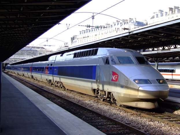 Hohe-Geschwindigkeitszug auf Schienen neben einem Bahnsteig mit Gebäuden und einem klaren blauen Himmel im Hintergrund.