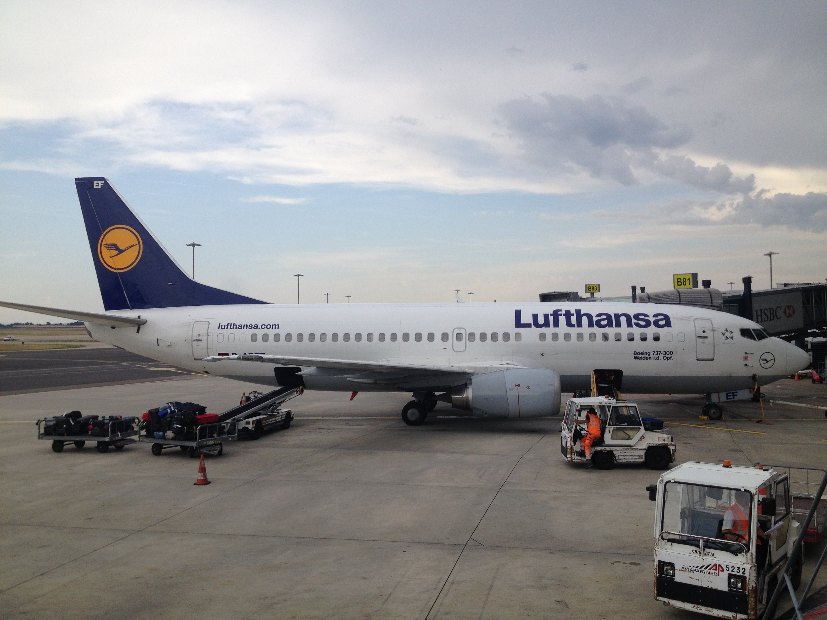 Airbus A320-200 der Lufthansa auf dem Frankfurter Flughafen geparkt mit Bodenfahrzeugen, Verkehrskegeln und Informationsschildern bei bewölktem Himmel.