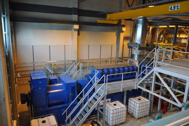 Ein Fabrikinnenraum mit Trägern, einer Treppe, Geländer, Containern, Lichtern, Rohren und Wänden.