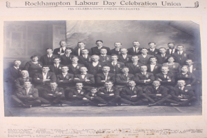 Eine Gruppe von Menschen in Blazern und Krawatten, die vor einer Wand mit der Aufschrift "Rockhampton Labour Day Celebration Union" posieren