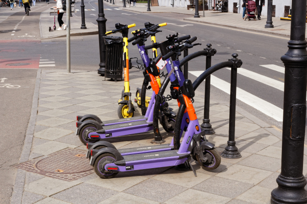Eine Reihe von Elektroscootern, die auf der Seite einer Straße geparkt sind, mit Menschen, die auf dem Gehweg gehen und Fahrräder fahren, einem Mülleimer im Vordergrund und Gebäuden, Bäumen und Pflanzen im Hintergrund.