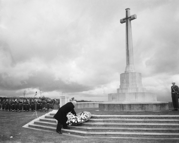 Ein Mann legt einen Kranz auf die Stufen eines kreuzförmigen Weltkriegsdenkmals, umgeben von einer Gruppe von Menschen, unter einem bewölkten Himmel mit Bäumen im Hintergrund, in Schwarz-Weiß.