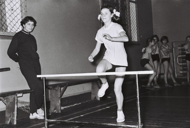 Schwarz-weißes Foto einer Frau beim Hürdenlauf in einer Halle mit Zuschauern und Fenstern im Hintergrund.