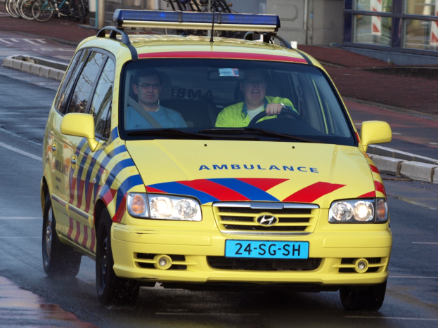 Ein gelber Krankenwagen mit zwei sichtbaren Insassen fährt an parkenden Fahrrädern auf einem Gehweg vorbei, mit einem Glasfenster-fassadengebäude im Hintergrund.
