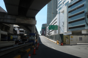 Stadtstraße mit Verkehrskegeln, Fahrzeugen, einer Brücke auf der linken Seite, Gebäuden mit Namensschildern, Laternenmästen und einem Himmel als Hintergrund.