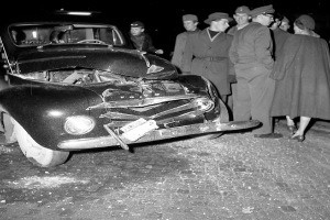 Eine schwarz-weisse Szene eines beschädigten Autos am Straßenrand, mit einer Gruppe von Menschen drumherum, die das Auto besorgt anschauen.