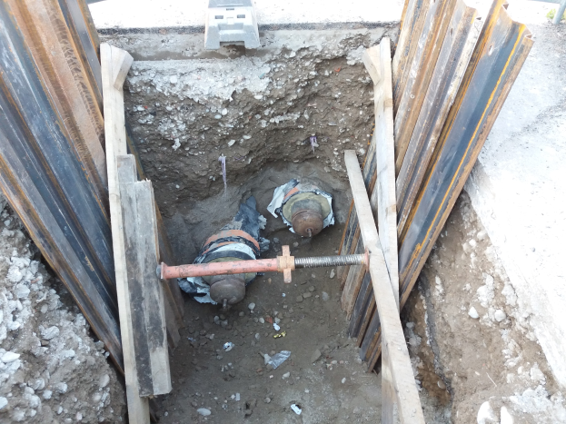 Baustelle mit einem gro├čen ausgehobenen Loch, Metallverstrebungen, verstreuten Steinen und einem Fahrzeug oben.
