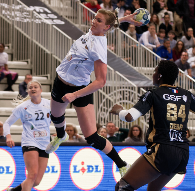 Eine Gruppe von Frauen, die auf einem Handballfeld Handball spielen, mit einem Ball in der Luft, Zuschauern auf Treppen und Geländern im Hintergrund und einem Schild mit der Aufschrift "Handball-Weltmeisterschaft 2019 - Tag 2".