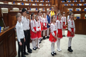 Eine Gruppe von Kindern in weißen und roten Kleidern steht zusammen in einem Raum, mit einem Podium und einem Mikrofon auf der linken Seite und Stühlen und Tischen mit verschiedenen Gegenständen im Hintergrund, die Flaggen tragen, die die Teilnahme an einer Nationalversammlung anzeigen.