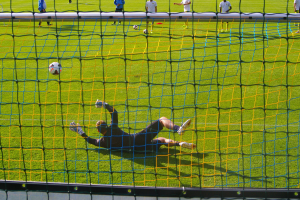Ein Fussballer taucht ab, um einen Ball vor einem Tor zu halten, mit einem Netz im Vordergrund und einigen Menschen im Hintergrund, während mehrere Bälle auf dem Boden verstreut sind.