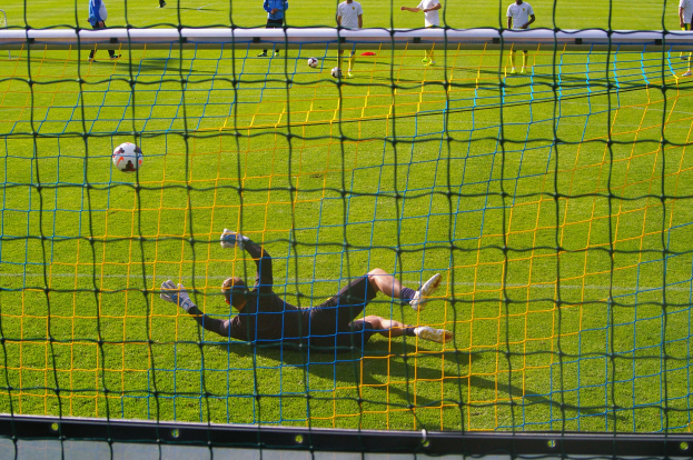 Ein Fussballer taucht ab, um einen Ball vor einem Tor zu halten, mit einem Netz im Vordergrund und einigen Menschen im Hintergrund, während mehrere Bälle auf dem Boden verstreut sind.