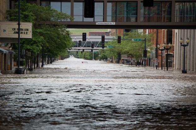 Überschwemmte Stadtstraße mit Wasser, das die Straße, die Infrastruktur und Gebäude bedeckt, einschließlich einer sichtbaren Brücke im Hintergrund.