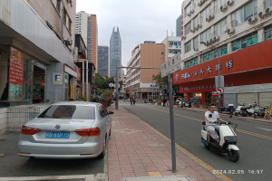 Ein Mann fährt mit einem Roller eine Straße entlang, die von hohen Gebäuden gesäumt ist. Menschen gehen auf den Gehwegen, Fahrzeuge fahren auf der Straße und Bäume unter einem klaren blauen Himmel sind im Hintergrund zu sehen.