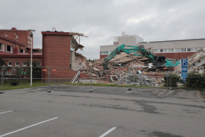 Großes Gebäude auf einem Parkplatz mit einem Bagger, Metallstangen, Zaun, Hinweisschild, Pflanzen, Gras, Bäumen, benachbarten Gebäuden und einem bewölkten Himmel abgebrochen.