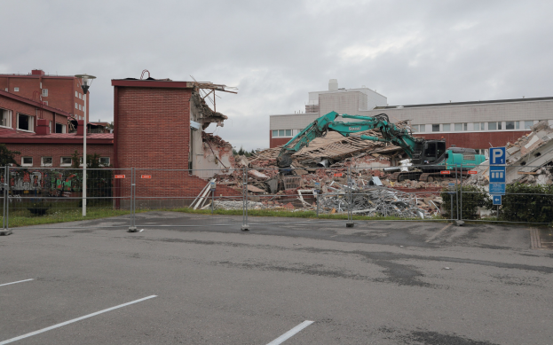 Großes Gebäude auf einem Parkplatz mit einem Bagger, Metallstangen, Zaun, Hinweisschild, Pflanzen, Gras, Bäumen, benachbarten Gebäuden und einem bewölkten Himmel abgebrochen.