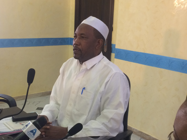 Ein Mann in weißem Hemd und Hut sitzt an einem Tisch mit Mikrofonen und spricht, während Papiere und Gegenstände auf dem Tisch sichtbar sind; rechts von ihm sitzt eine Person mit Brille, im Hintergrund eine Tür und eine Wand.