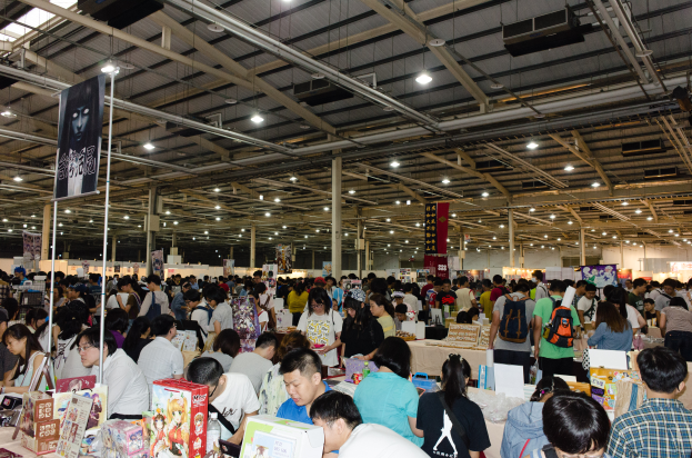 Eine große Gruppe von Menschen umringt Tische, die mit Büchern, Kartons und anderen Gegenständen bedeckt sind, auf einer Buchmesse, mit Schildern und Lichtern darüber.