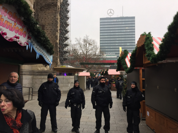 Eine Gruppe von Polizisten in schwarzen Uniformen und Mützen steht vor einem Weihnachtsmarkt in Berlin, Deutschland, mit festlichen Dekorationen, Gebäuden, Bäumen und einem klaren blauen Himmel im Hintergrund.