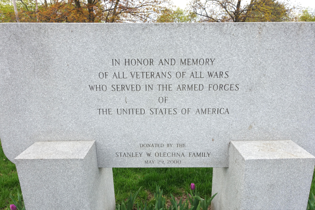 Ein Gedenkstein mit einer Inschrift, die alle US-Veteranen ehrt, umgeben von Pflanzen, Blumen, Gras, Bäumen und unter einem bewölkten Himmel.