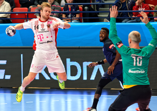 Eine Gruppe von Männern, die auf einem Handballfeld mit einem Ball in der Mitte spielen, während Zuschauer von Stühlen und Geländern im Hintergrund zusehen, mit einer Tafel, auf der "Futsal-Weltmeisterschaft 2019 - Bayern München vs. Paris Saint-Germain" steht.