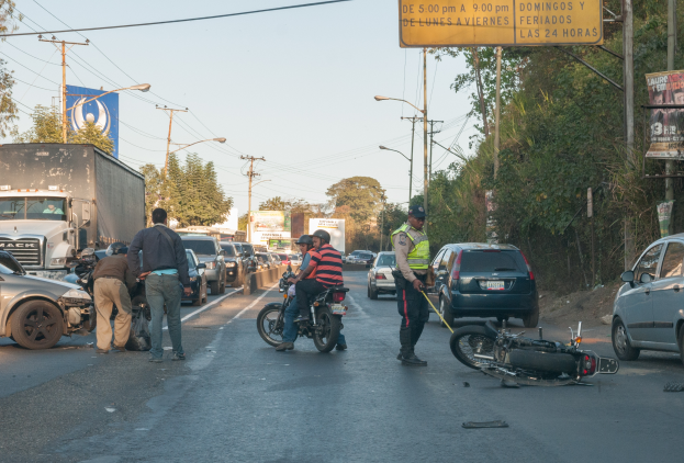 Eine Gruppe von Menschen umringt ein verunglücktes Motorrad am Straßenrand mit mehreren Fahrzeugen, darunter ein Lastwagen, und einer Hintergrundlandschaft aus Bäumen, Pfählen, Laternen und Schildern unter dem Himmel.