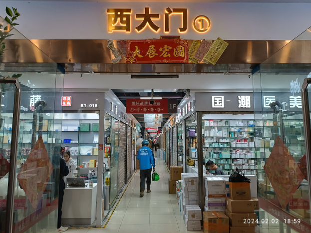 Innenraum einer chinesischen Apotheke mit Glaswänden, einer Pflanze auf der linken Seite, Regalen mit Gegenständen, verstreuten Kartons und einem Namensschild oben.