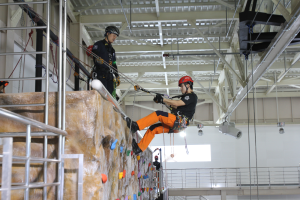 Zwei Individuals in schwarzen und orangen Outfits und Helmen klettern an einer Felswand in einer Halle, gesichert durch Seile, mit einem Metallgeländer links und einer weißen Wand mit Lampen im Hintergrund.