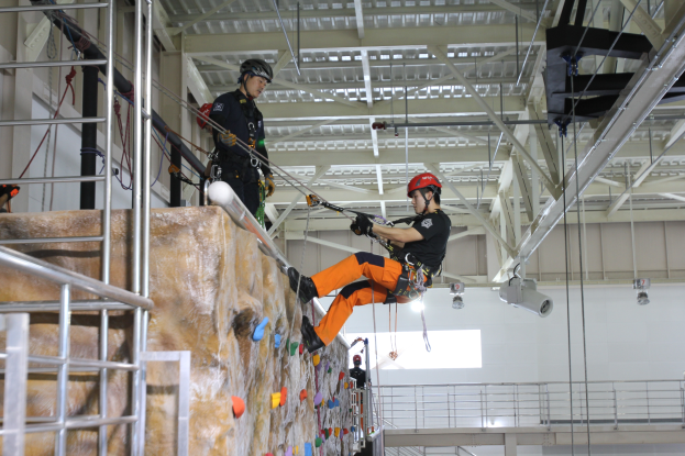Zwei Individuals in schwarzen und orangen Outfits und Helmen klettern an einer Felswand in einer Halle, gesichert durch Seile, mit einem Metallgeländer links und einer weißen Wand mit Lampen im Hintergrund.