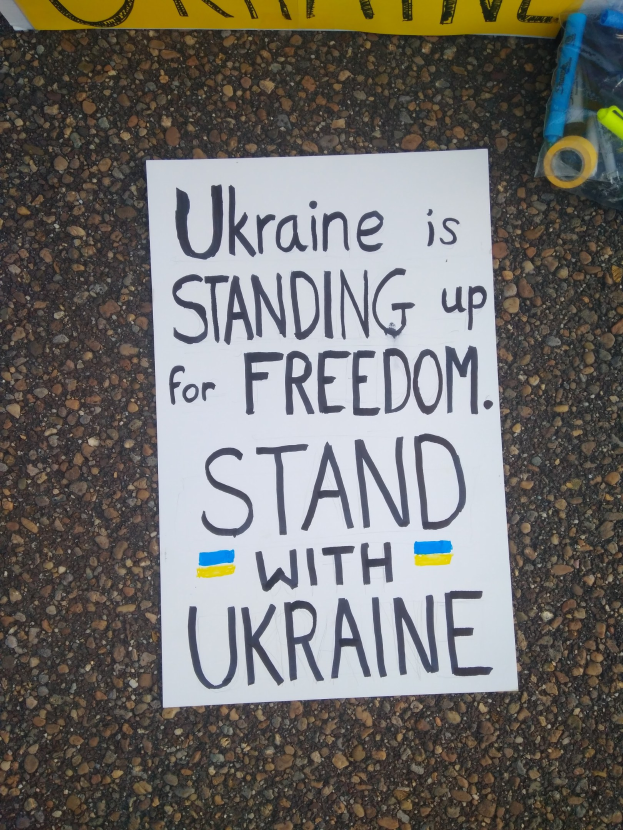 Weißes Papier mit dem Text "Ukraine steht für Freiheit ein, steht mit Ukraine" auf dem Boden vor einem gelben Banner, mit einer Plastikhülle mit verschiedenen Gegenständen rechts.