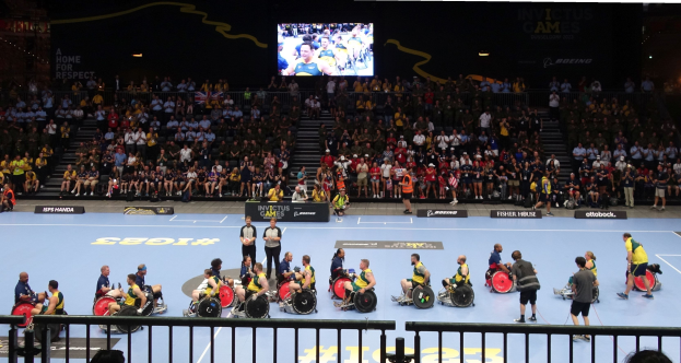 Eine Gruppe von Rollstuhlfahrern bei den Invictus Games Düsseldorf 2020 auf einem Court umgeben von einem Geländer, mit einer großen Menge im Hintergrund und Stufen, die zu einem Bildschirm führen.