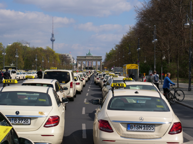 Eine lange Reihe von Taxis steht entlang einer vielbefahrenen Straße in Berlin, Deutschland, mit fahrenden Fahrzeugen, Fößlern und Radfahrern auf dem Gehweg, flankiert von Laternenmästen und Bäumen und Gebäuden, einem Bogen und einem Turm im Hintergrund unter einem bewölkten Himmel.