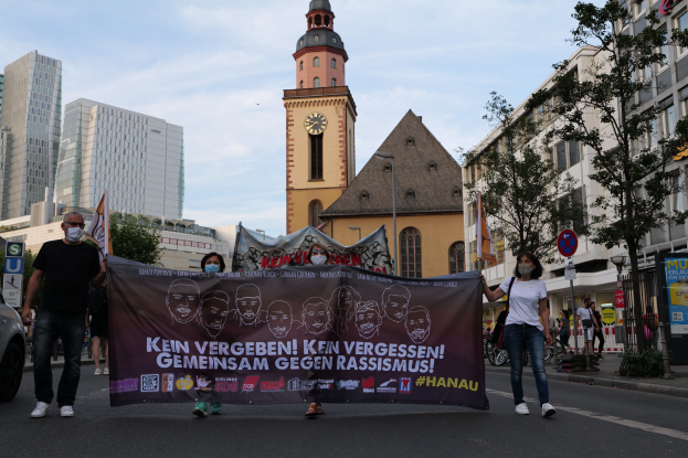 Eine Gruppe von Menschen in Masken, die eine Straße entlanggehen und eine Fahne halten, mit einem geparkten Auto auf der linken Seite, Gebäuden und Bäumen im Hintergrund und einem klaren blauen Himmel.