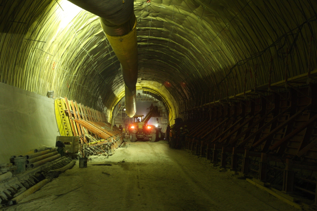 Baustelle mit einem großen zentralen Tunnel, Fahrzeugen, verstreuten hölzernen Gegenständen, Rohren und einer Wand auf der linken Seite, beleuchtet von Deckenlampen.