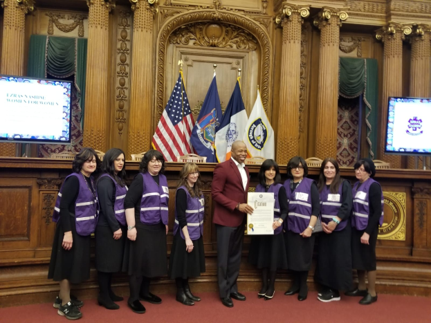 Gruppe von Menschen vor Flaggen mit einem Mann, der ein Zertifikat hält, flankiert von zwei Bildschirmen, vor einer hölzernen Wand mit Schnitzereien und Vorhängen, was auf eine Stadtratssitzung hinweist.