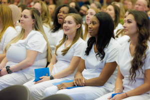Eine Gruppe von Frauen in weißen Kleidern sitzt in einem Hörsaal, jede hält ein blaues Objekt, mit einem unscharfen Hintergrund, der den Fokus auf sie legt, was darauf hindeutet, dass sie Teil eines Pflegeprogramms sind.