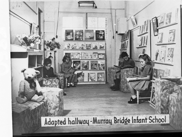 Eine Gruppe von Kindern sitzt auf Stühlen in einem Raum mit Bücherregalen, einem Teddybär, einer Blumenvase auf einem Tisch und gerahmten Bildern an der Wand, mit Text unten lesend "Angepasster Flur Murray Bridge Kindergarten"