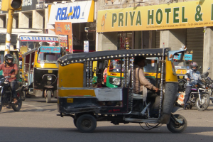 Eine belebte Straße mit zahlreichen Fahrzeugen, darunter ein Auto-Rikscha, und Menschen mit Helmen; eine Ampel mit einem Mast im Vordergrund; Gebäude mit Namensschildern und Rollläden im Hintergrund.