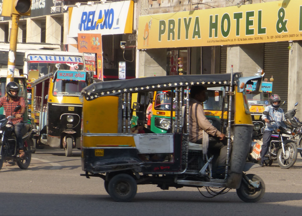 Eine belebte Straße mit zahlreichen Fahrzeugen, darunter ein Auto-Rikscha, und Menschen mit Helmen; eine Ampel mit einem Mast im Vordergrund; Gebäude mit Namensschildern und Rollläden im Hintergrund.