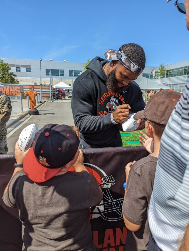 Cleveland Browns Wide Receiver Odell Beckham Jr. signiert Autogramme für Fans auf dem Trainingsgelände, umgeben von Menschen mit Kappen, einem Banner und Gebäuden im Hintergrund.
