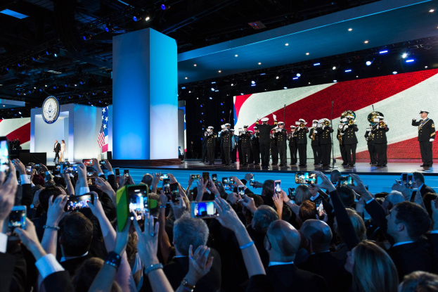 Eine Gruppe von Menschen auf einer Bühne, einige halten Telefone, mit einer Person auf einem Podium links, Flaggen, einem Bildschirm, einer Säule, einer Uhr und Deckenleuchten im Hintergrund, während einer Präsidentenrede.