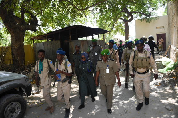 Eine Gruppe von Polizisten in Uniform, einige mit Waffen, die eine Straße entlanggehen neben einem Auto, mit Gebäuden, Bäumen und Schuppen im Hintergrund unter einem sichtbaren Himmel.