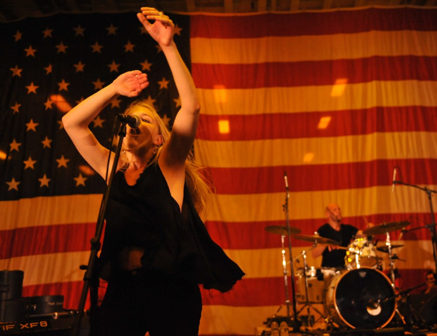 Eine Frau singt in ein Mikrofon auf der Bühne mit einer amerikanischen Flagge im Hintergrund, begleitet von einem Schlagzeuger, mit Bühnenausrüstung wie Mikros, Ständer, Lautsprecher und Beleuchtung.