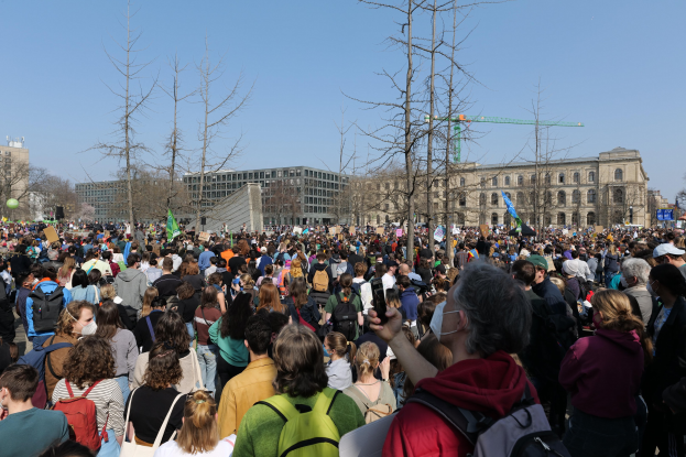 Eine große Menschenmenge steht vor einem Gebäude mit Fenstern, Bäumen und einem klaren Himmel, viele halten Schilder und tragen Taschen, was auf eine Klimaprotest hingedeutet.
