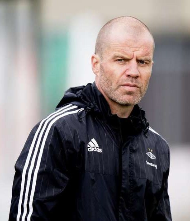 Baldiger Trainer in marineblauer Jacke mit weißen Streifen und weißem Kragen auf einem Fußballfeld stehend, Hände verschränkt, mit ernster Miene.