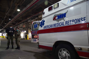 American Medical Response ambulance parked indoors with two masked individuals in front of it, a vehicle to the left, and additional people and equipment in the background.
