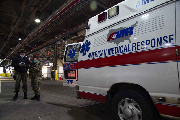American Medical Response ambulance parked indoors with two masked individuals in front of it, a vehicle to the left, and additional people and equipment in the background.