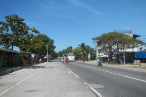 Stadtstraße mit fahrenden Autos, Fußgängern auf dem Gehweg, Geschäftsfronten, Bäumen, Gebäuden, Stromleitungen an Masten und einem bewölkten Himmel.